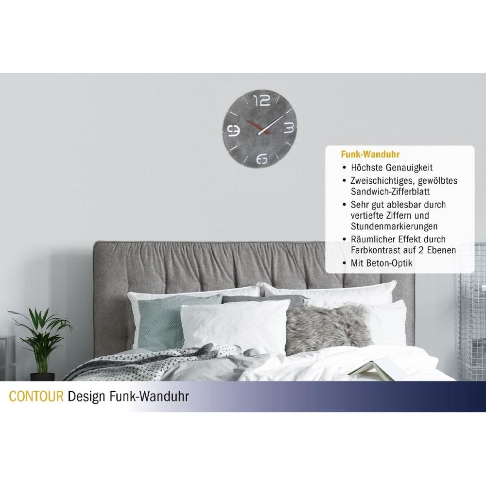 Graue Wanduhr mit Betonoptik über einem Bett mit weißen Kissen.