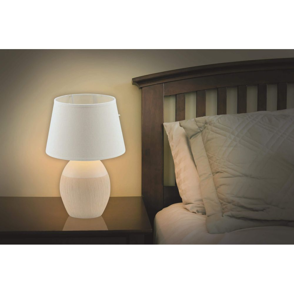 Eine Nachttischlampe mit weißem Schirm steht neben einem Bett mit dunklem Holz.