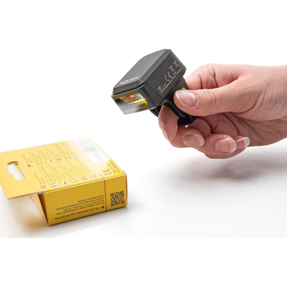 Una mano tiene uno scanner di codici a barre ad anello nero puntato verso una scatola gialla.