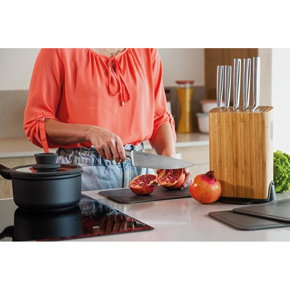 Een vrouw snijdt granaatappels in een keuken. Een mes, snijplank, pan en messenset zijn zichtbaar.