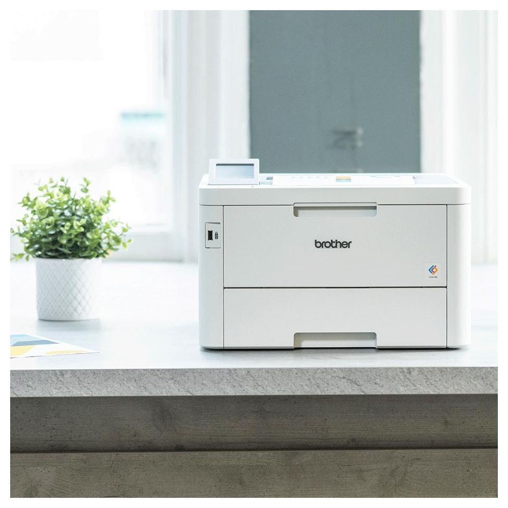Une imprimante Brother blanche est posée sur un bureau avec une plante verte à sa gauche.
