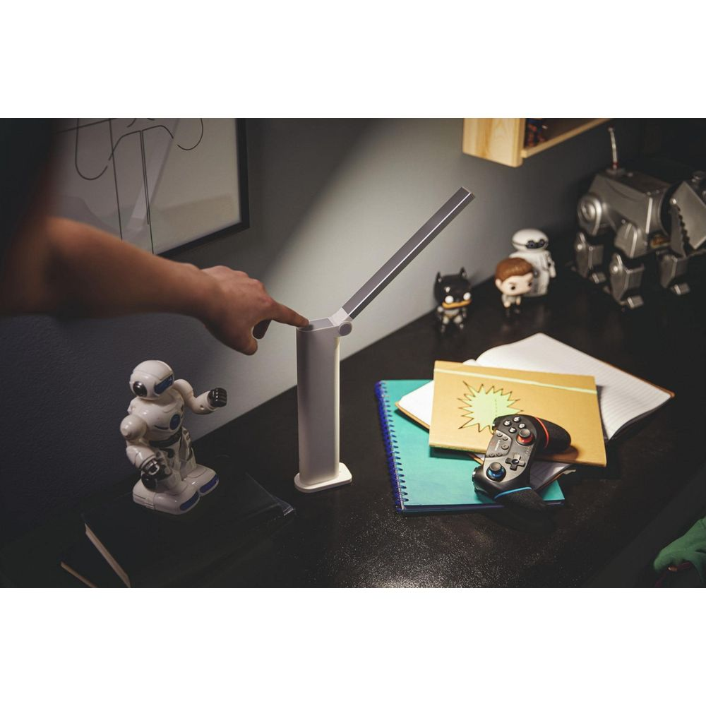 Ein Finger berührt eine weiße Lampe auf einem Schreibtisch mit Spielzeugrobotern.
