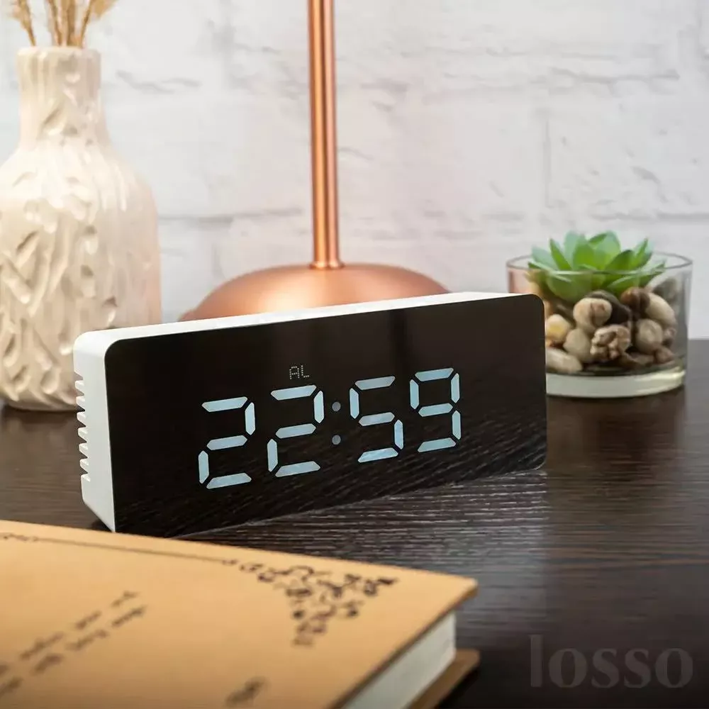 Cyfrowy zegar pokazuje 22:59 na stoliku nocnym obok wazonu i rośliny.