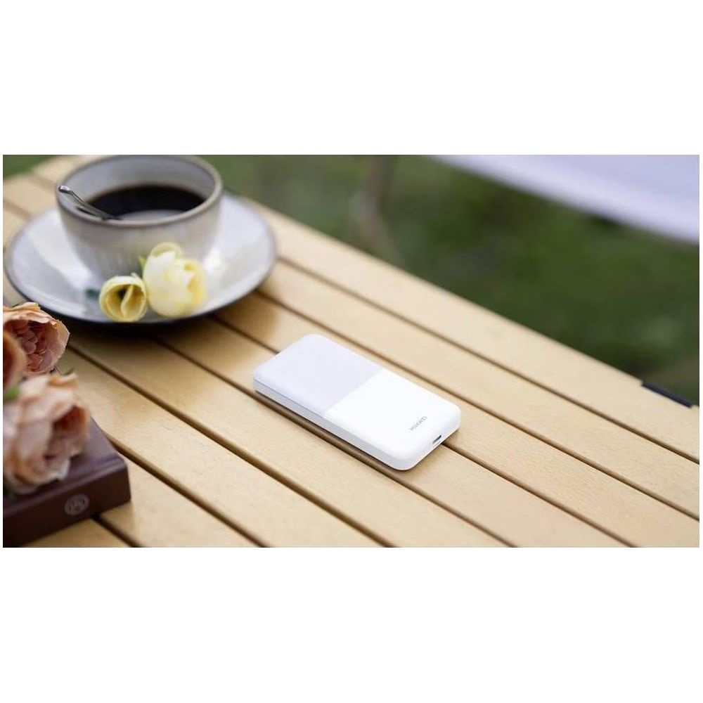 Biały powerbank leży na drewnianym stole obok filiżanki kawy i kwiatów.