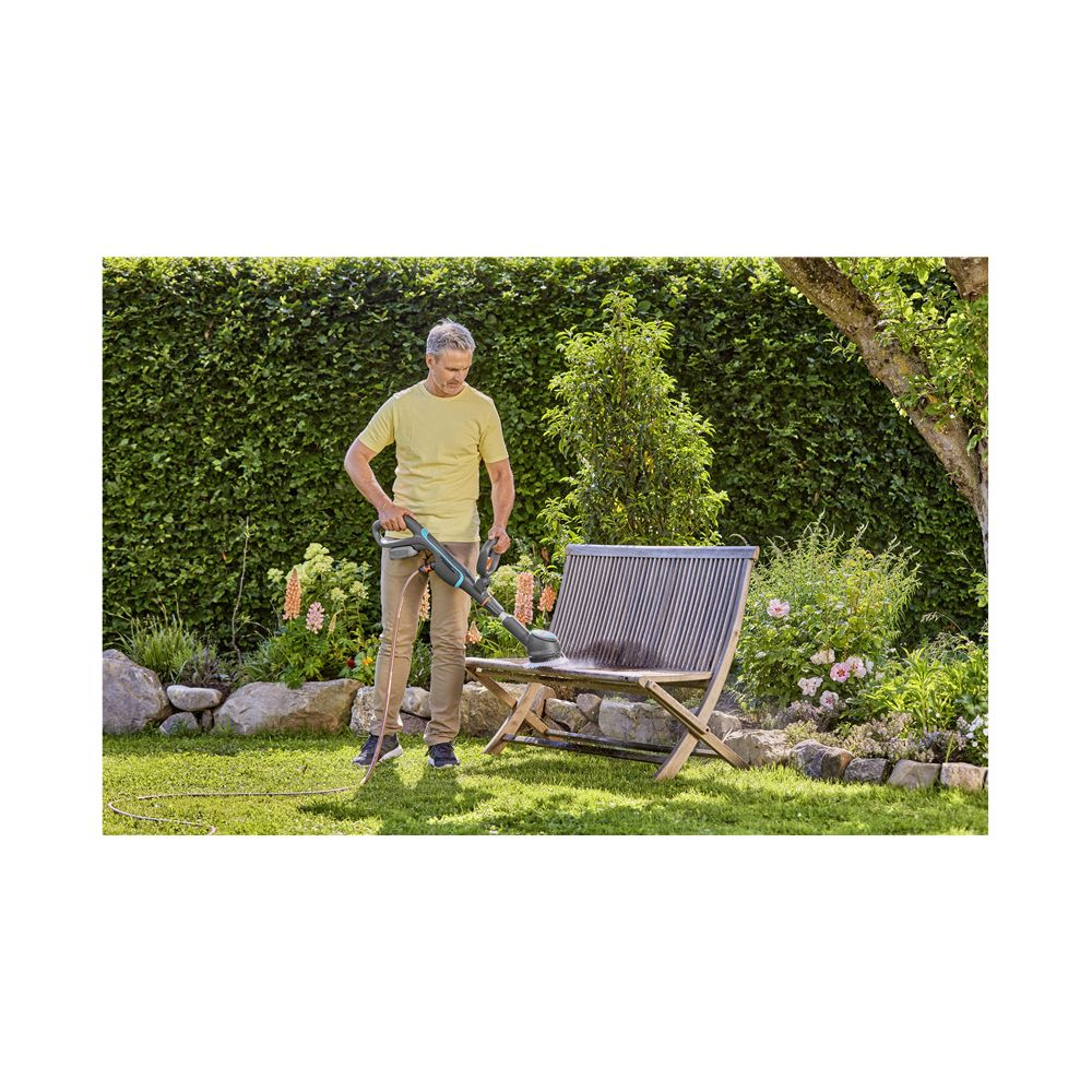 Mann reinigt Gartenbank mit Gerät, grüne Hecke im Hintergrund.