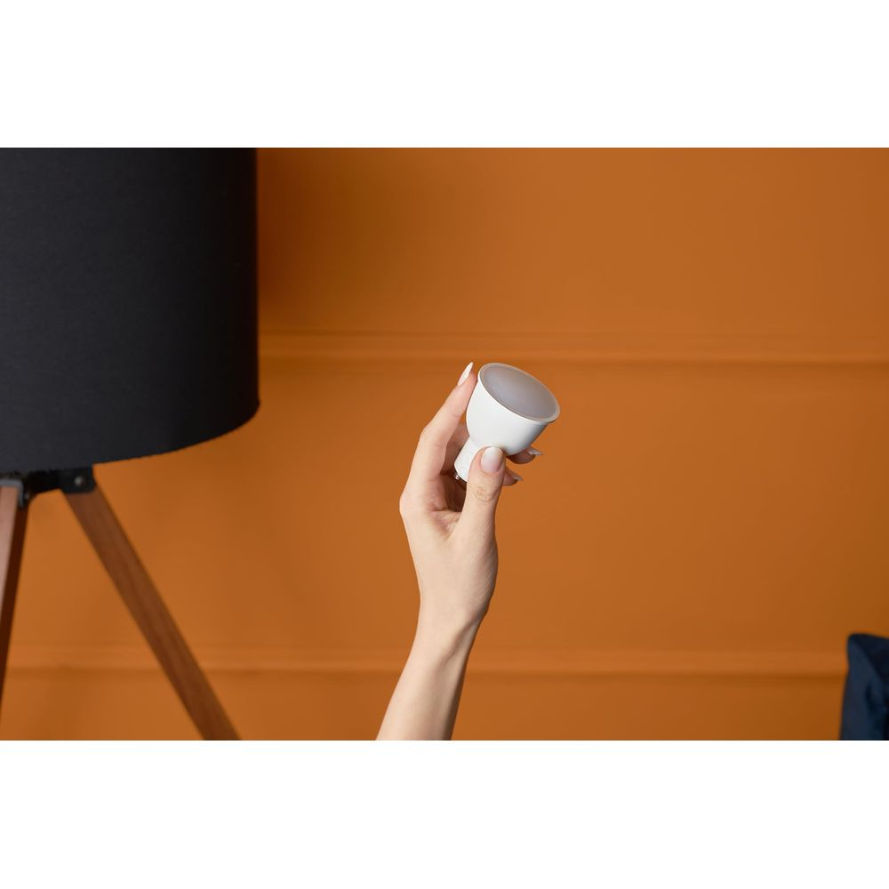 Una mano tiene una lampadina LED bianca contro un muro arancione, vicino a un paralume nero.