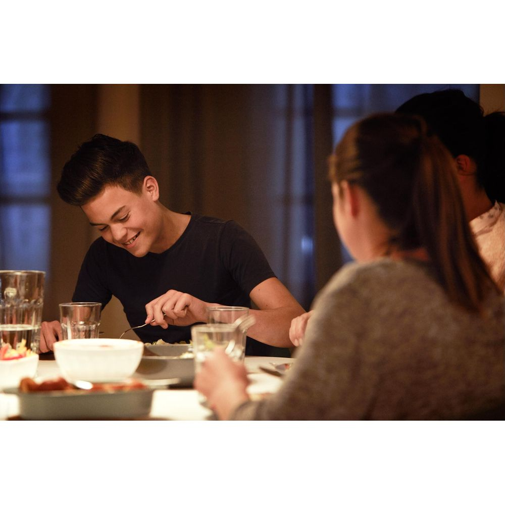 Młoda osoba je przy stole z rodziną, uśmiecha się i patrzy na talerz.