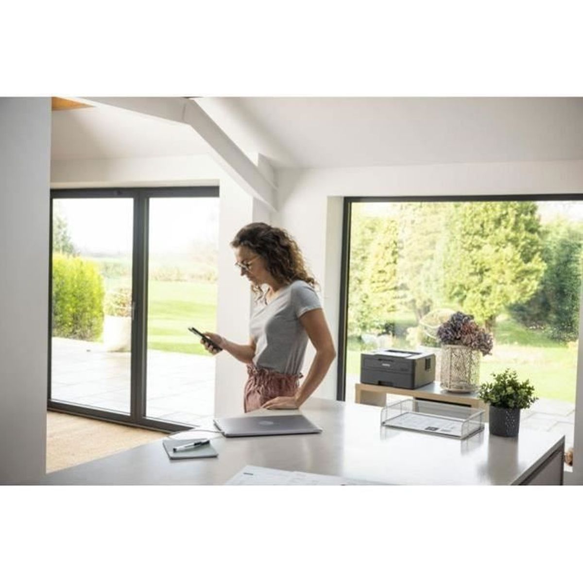 Kobieta z telefonem przy biurku z laptopem, drukarką i roślinami.