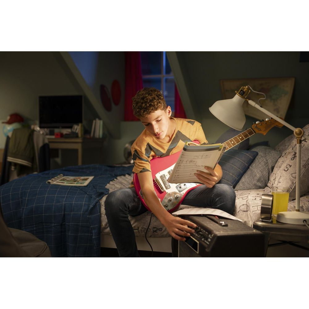 Een jongen zit op een bed, speelt een rode gitaar en leest bladmuziek.