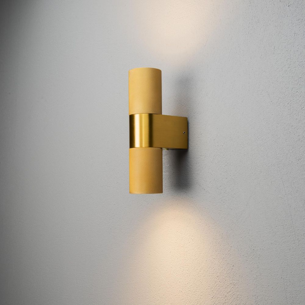 Eine goldene Wandleuchte mit zwei zylindrischen Lampenschirmen an einer grauen Wand.