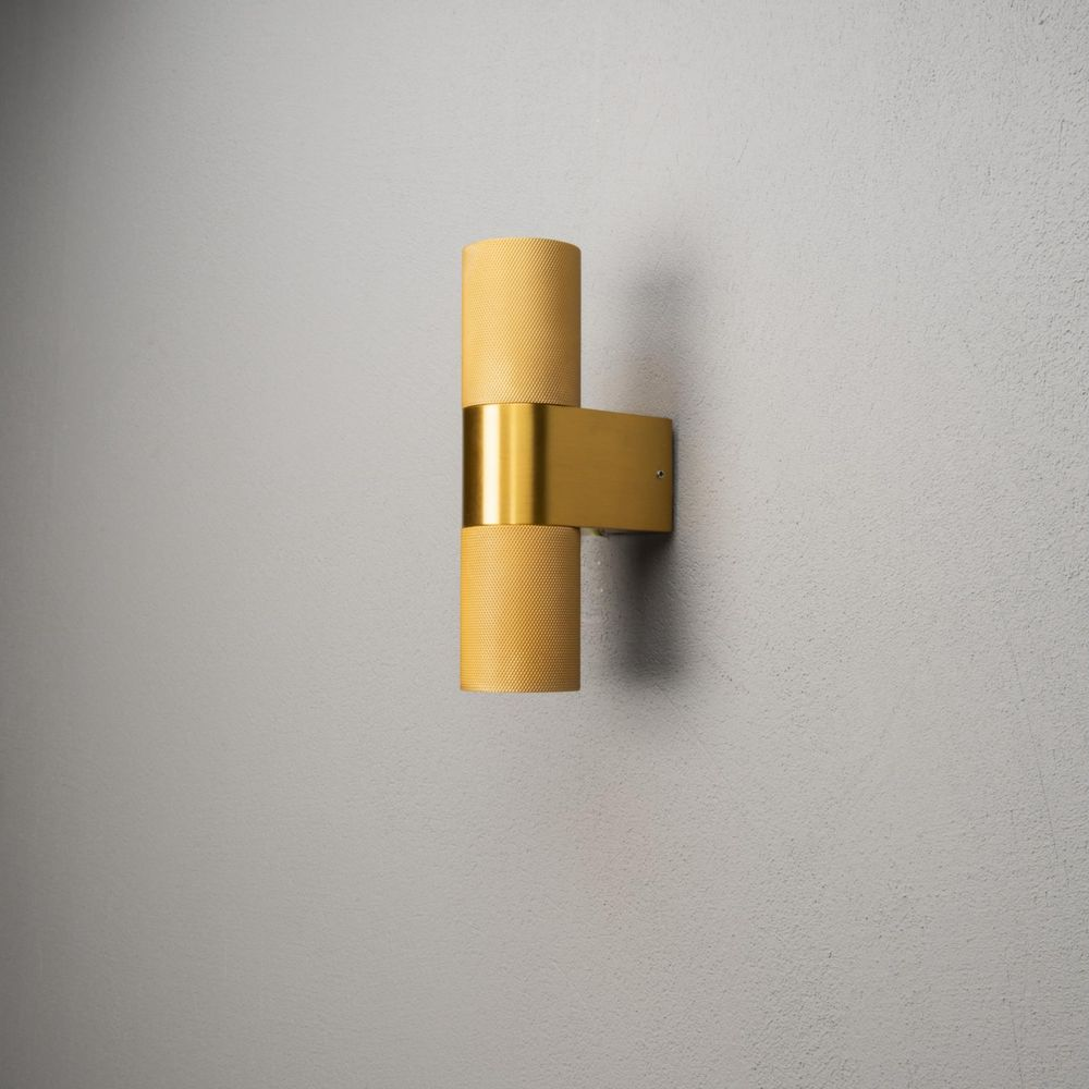 Eine goldene Wandleuchte mit zwei zylindrischen Lampenschirmen an einer grauen Wand.