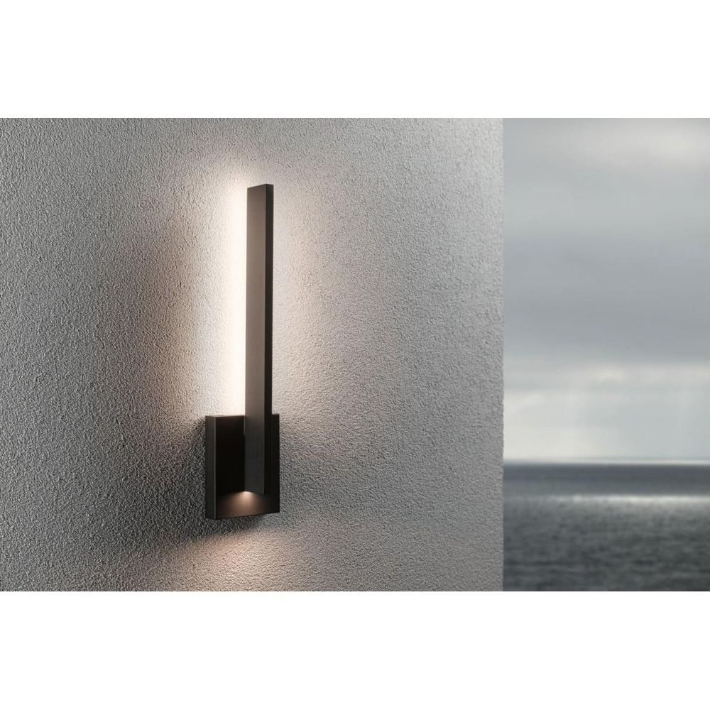 Nowoczesna czarna lampa ścienna oświetla teksturowaną ścianę. Po prawej morze pod pochmurnym niebem.