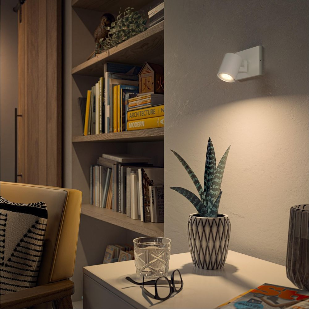 Een witte wandspot verlicht een sanseveria op een tafel naast een boekenplank.