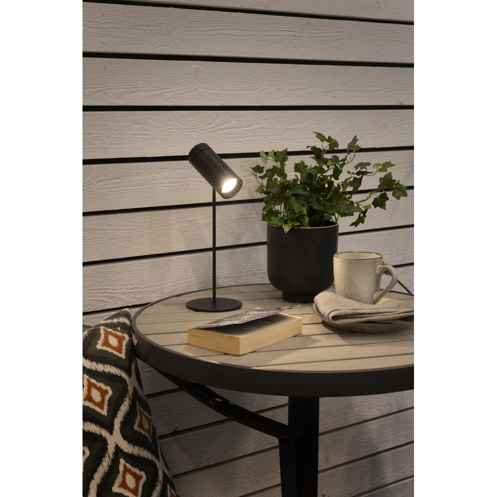 Ein schwarzer Tisch mit Lampe, Buch, Topfpflanze, Tasse und Kissen vor Holzwand.