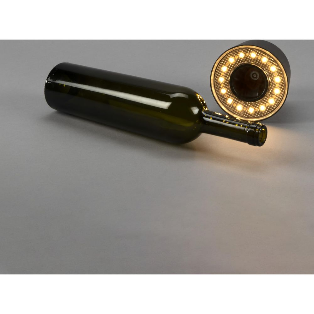 Eine dunkle Glasflasche liegt auf einem grauen Hintergrund neben einer runden LED-Leuchte.