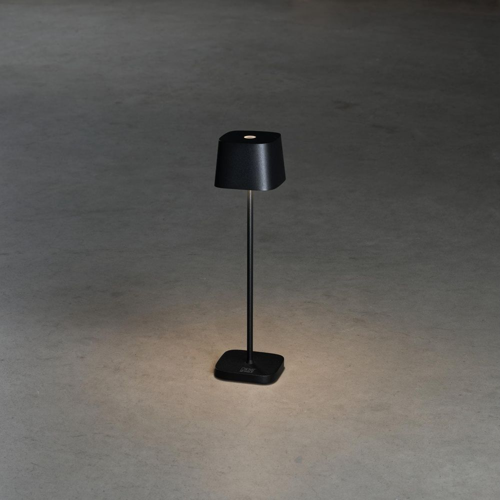 Una lampada da tavolo nera con paralume cilindrico poggia su una base rettangolare.