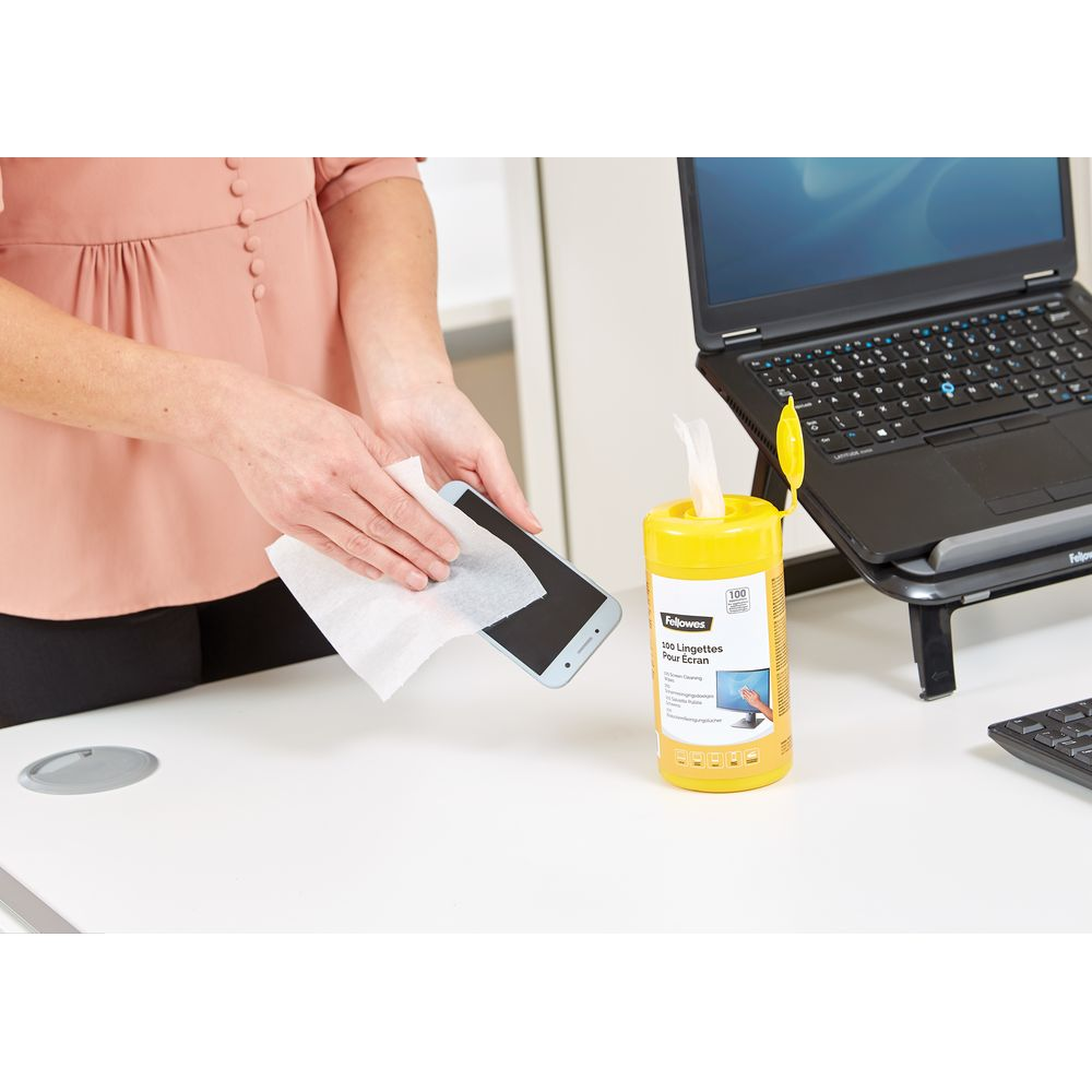Persona pulisce smartphone con salviettina accanto a laptop e contenitore salviettine.