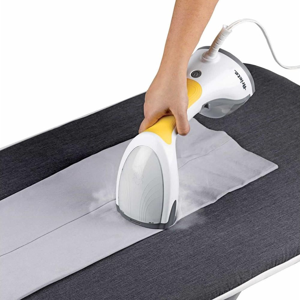 Dłoń trzyma biało-żółtą parownicę do ubrań nad szarą tkaniną na desce do prasowania.