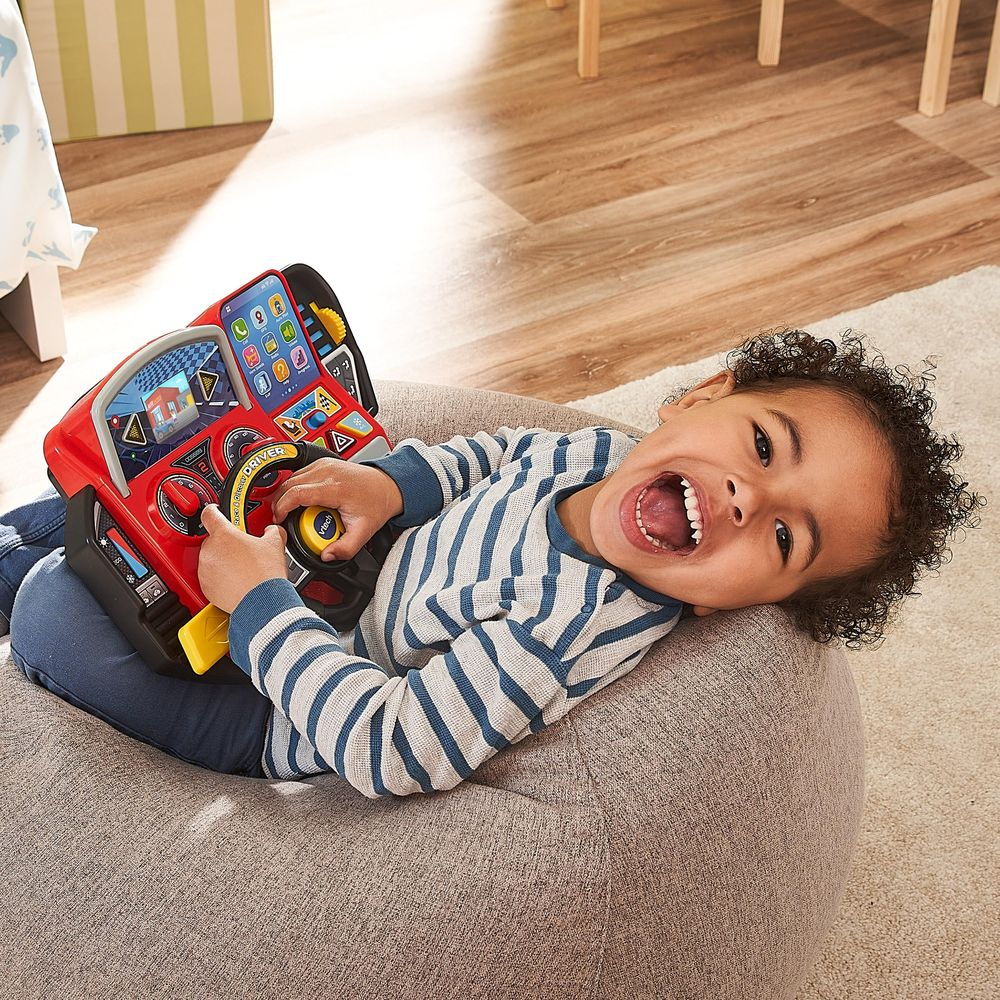 Un enfant souriant est allongé sur un pouf, tenant un volant de jouet rouge.