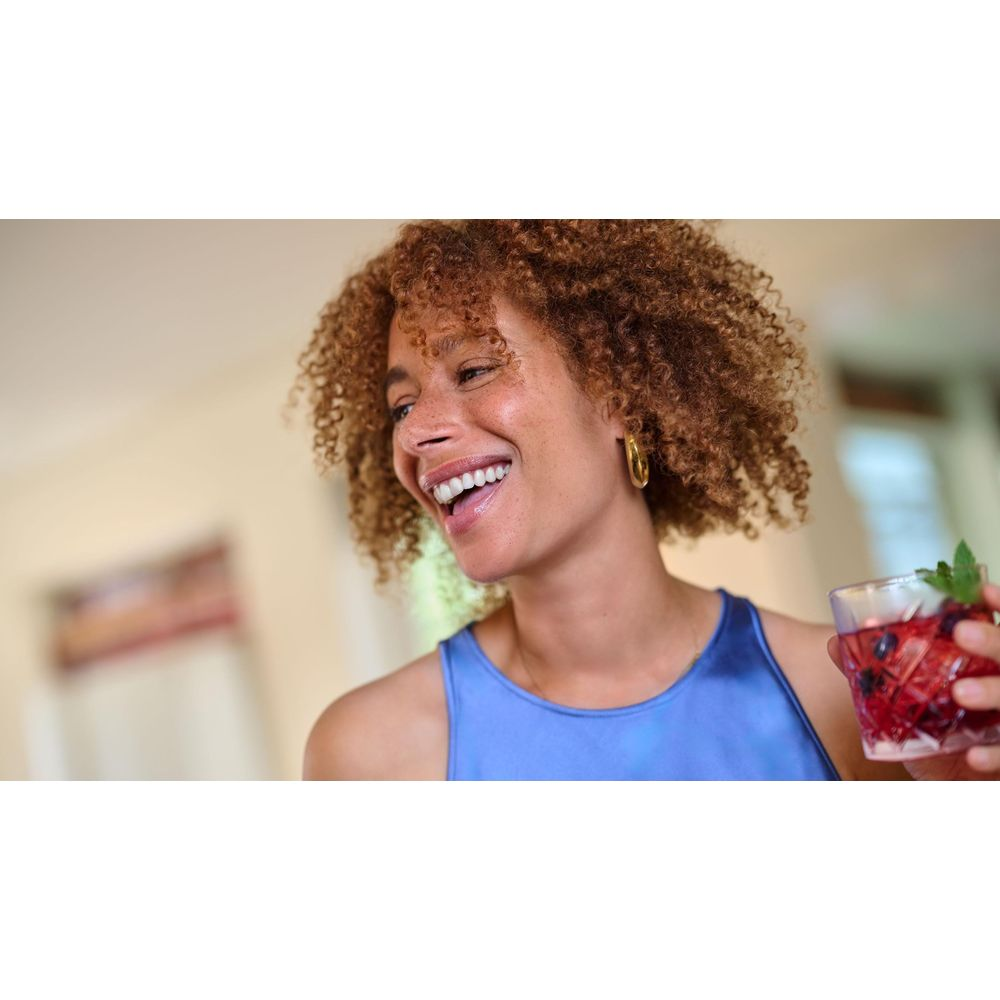 Frau mit lockigem Haar lacht und hält ein Glas mit Beeren und Minze.