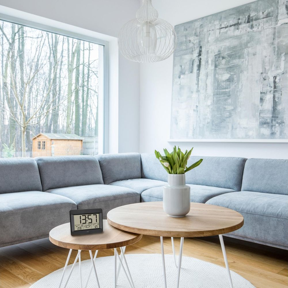 Ein modernes Wohnzimmer mit einem grauen Ecksofa, zwei Holztischen und einer Digitaluhr.