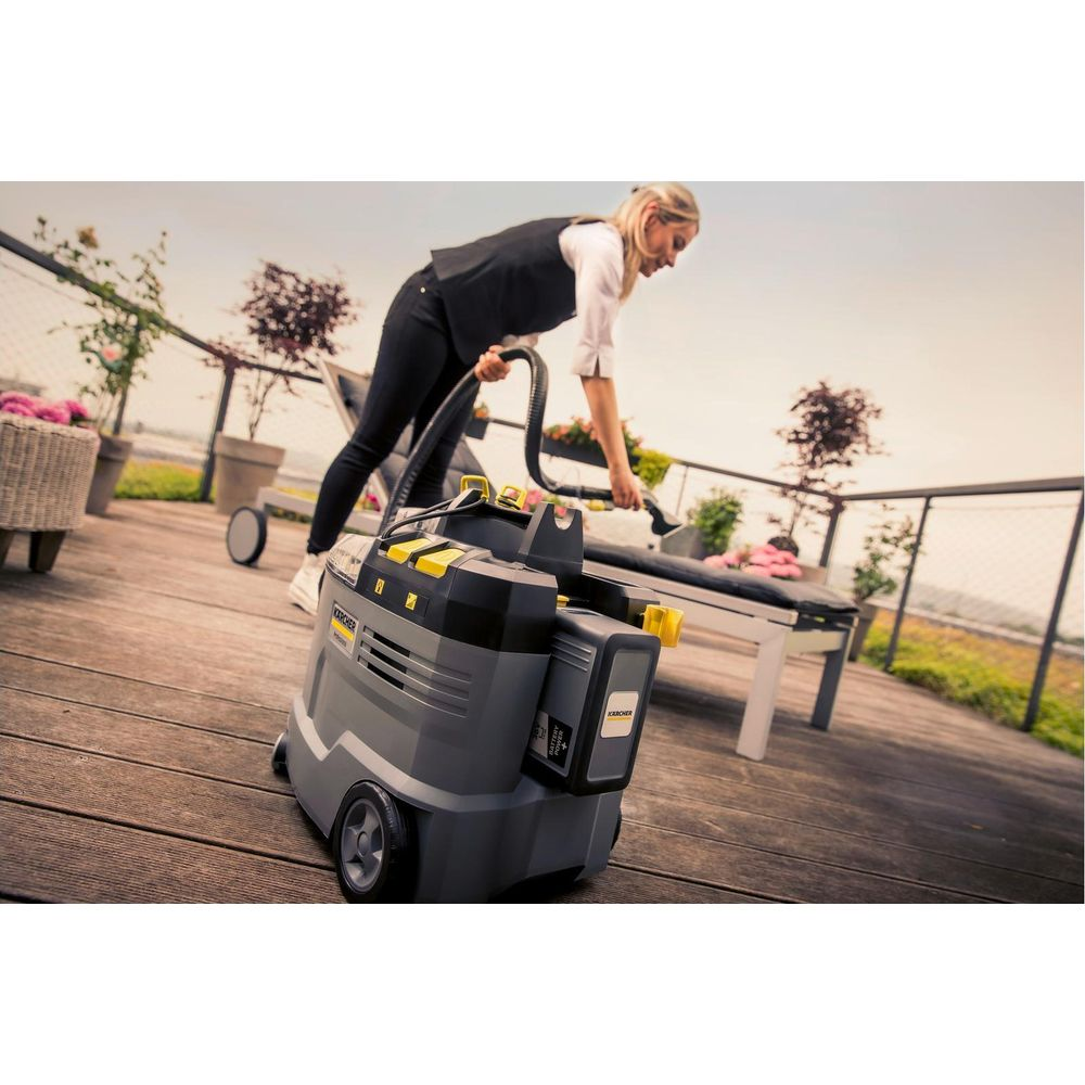 Eine Frau reinigt mit einem Kärcher-Sauger eine Gartenbank auf einer Holzterrasse.