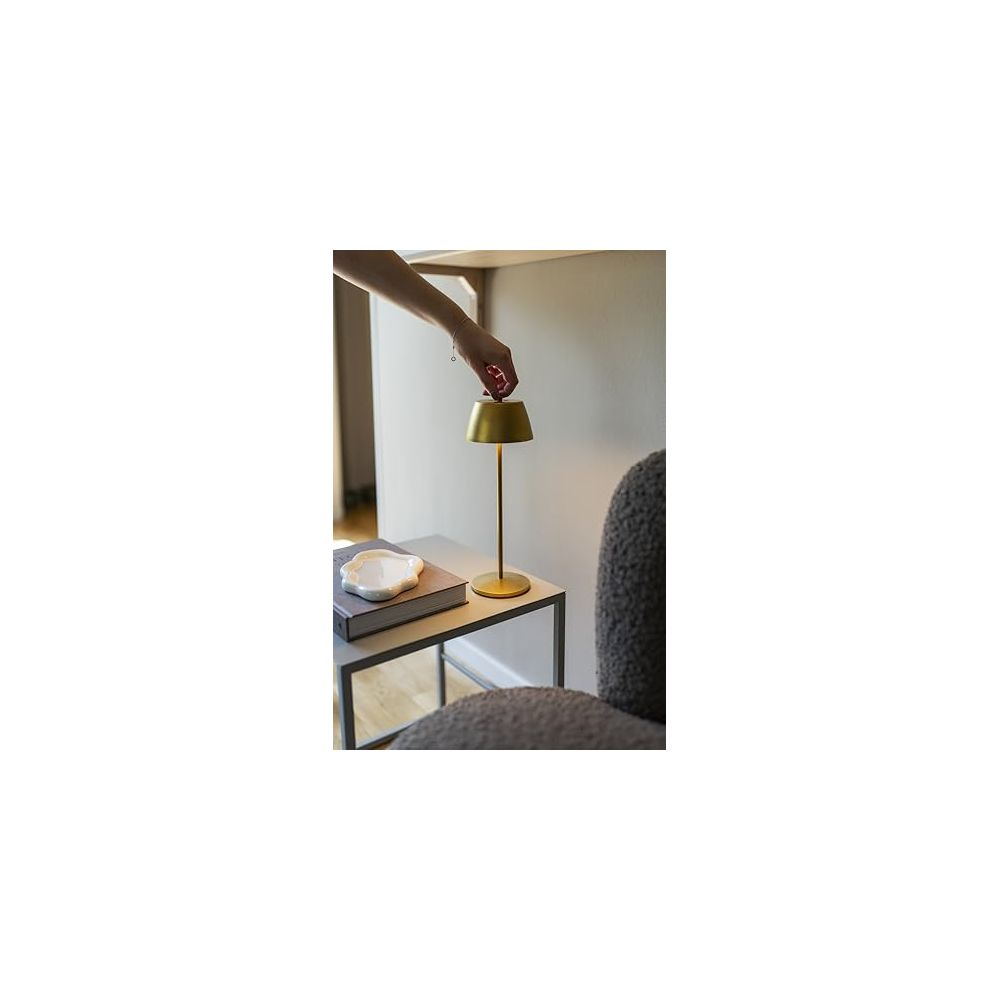 Eine Hand berührt eine goldene Lampe auf einem Beistelltisch neben einem Buch.