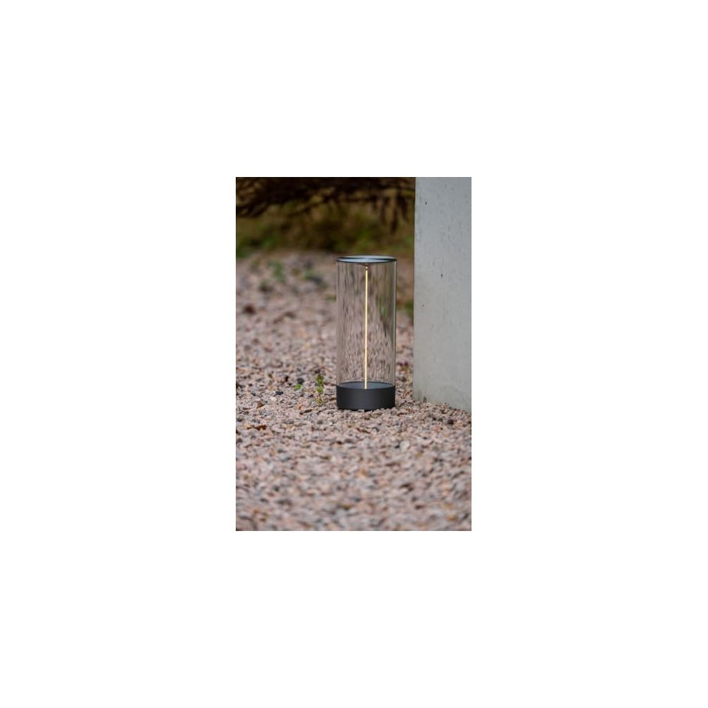 Eine zylindrische Lampe mit einem dunklen Sockel und Glaszylinder, die auf Kies steht.