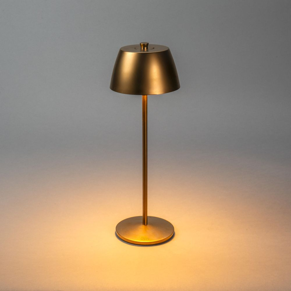 Una lampada da tavolo dorata con base rotonda e paralume conico su sfondo grigio.