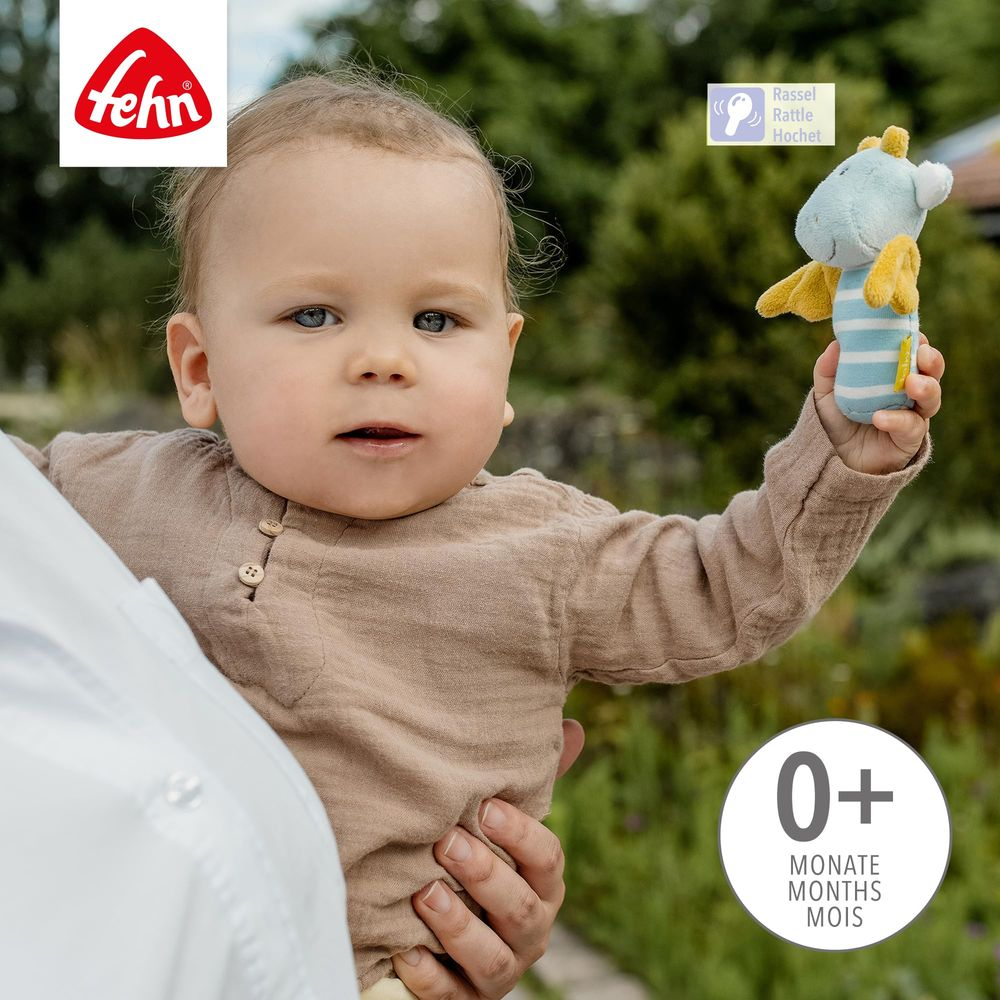 Un bébé tient un hochet serpent bleu avec des ailes jaunes. Logo Fehn en haut à gauche.