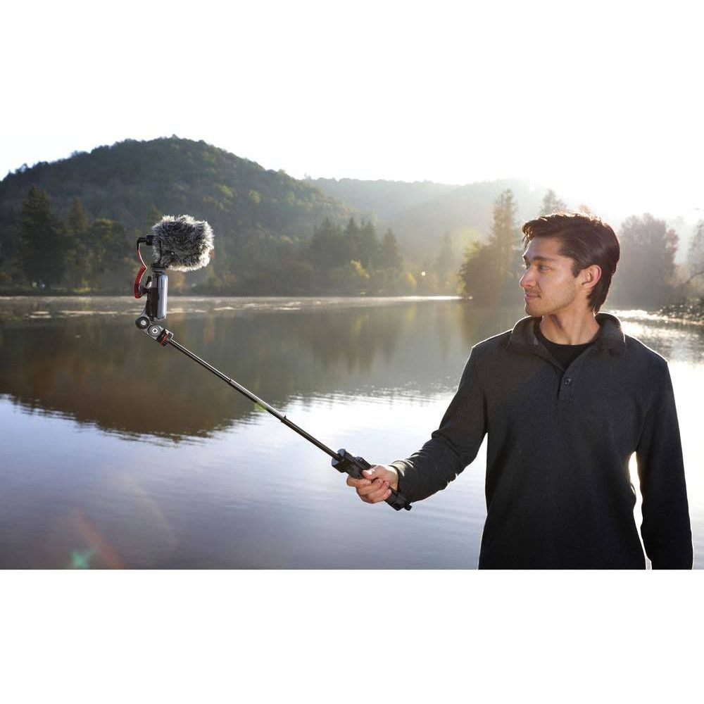 Man holds microphone pole over lake with tree-covered hills in background.