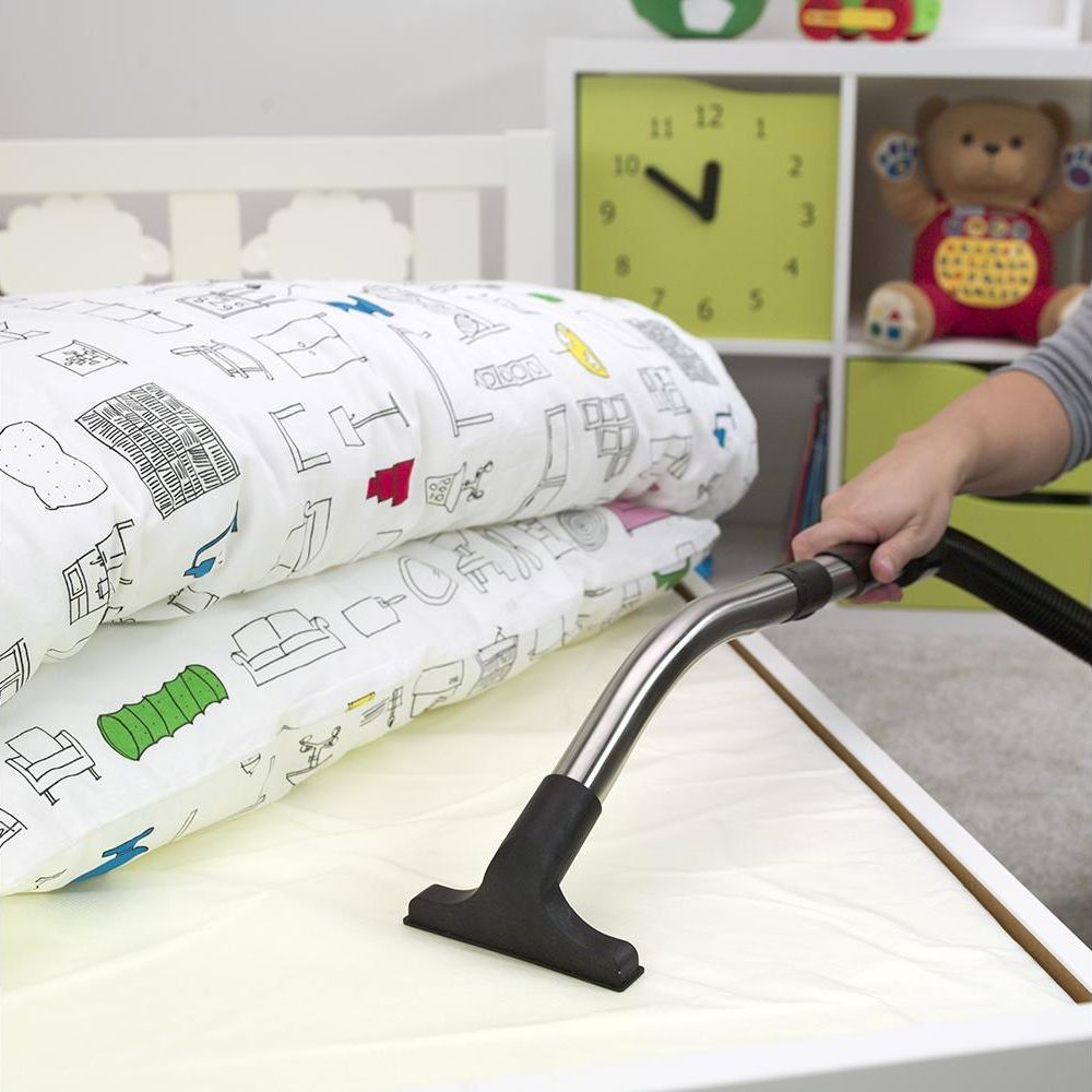 Una mano usa un aspirapolvere su un letto con biancheria da letto bianca. Giocattoli visibili.
