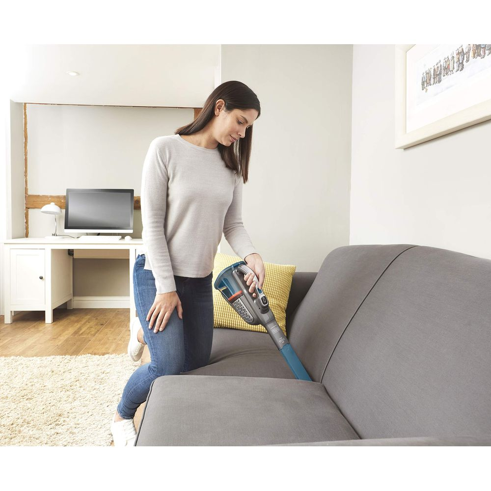 Frau saugt Sofa mit blauem Handstaubsauger. Innenraum mit Schreibtisch.