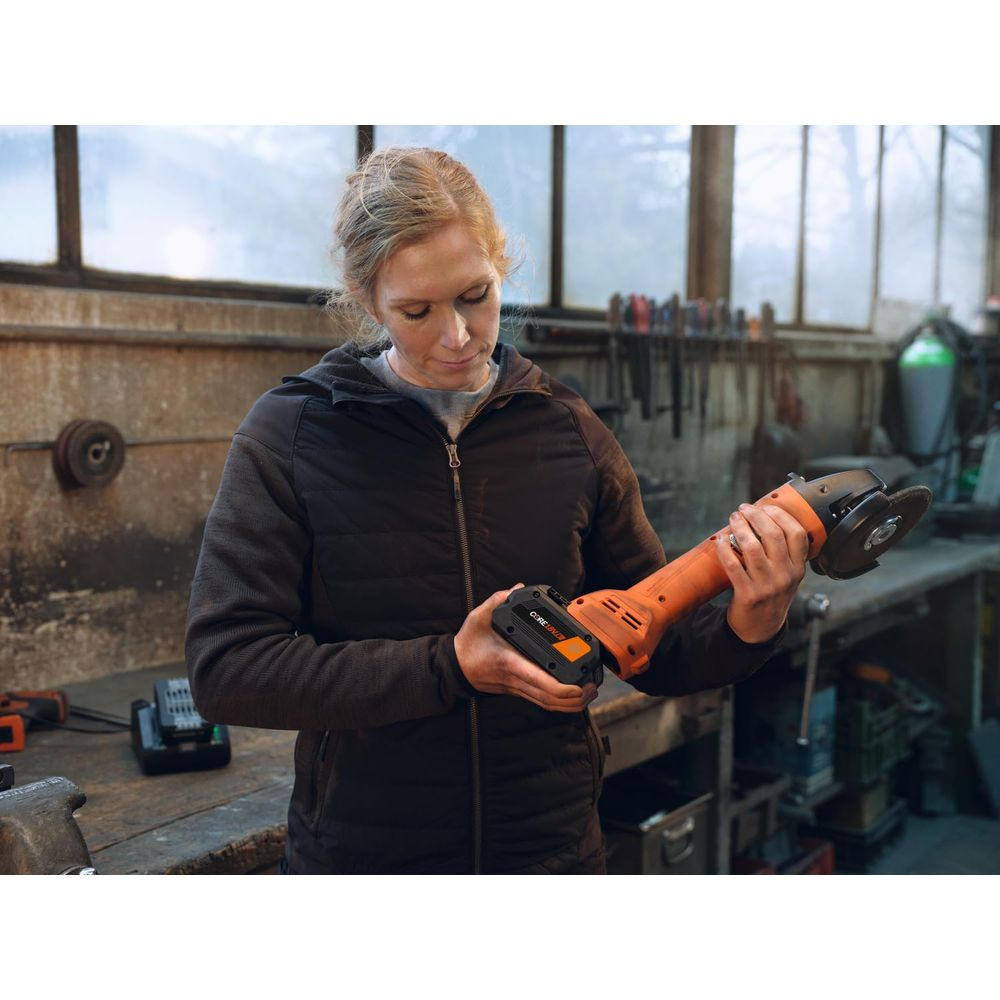 Eine Frau hält einen orangefarbenen Winkelschleifer in einer Werkstatt.