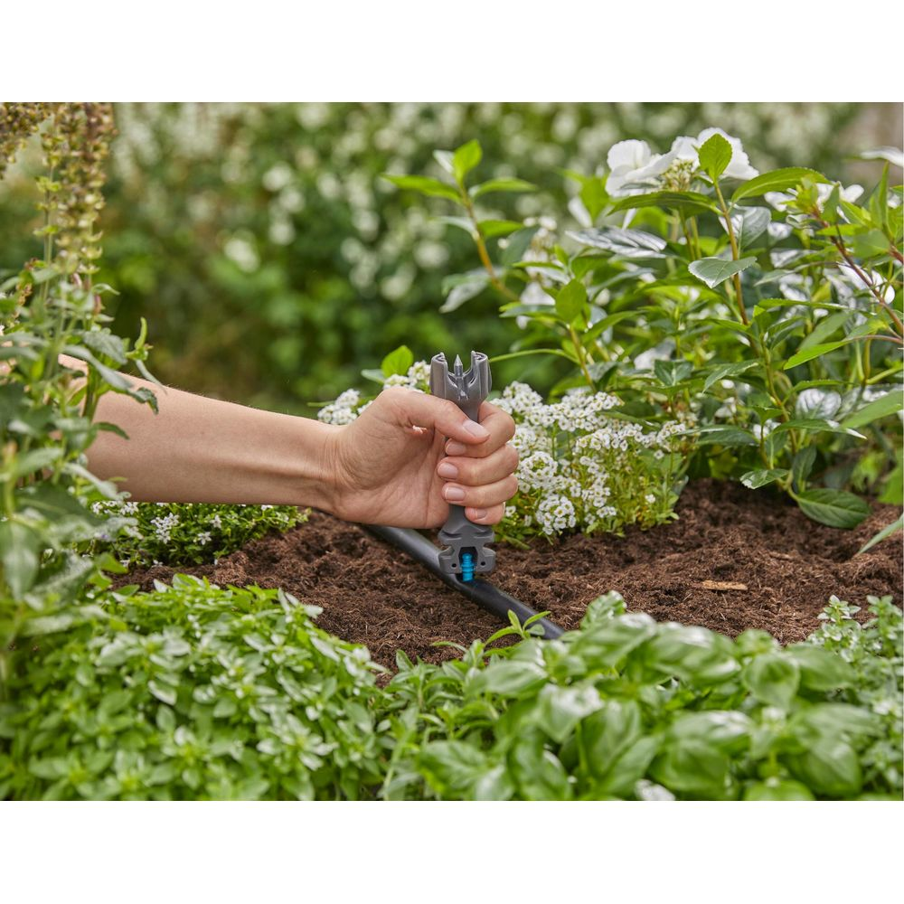 Een hand houdt een gereedschap boven een druppelirrigatieslang in een tuin.