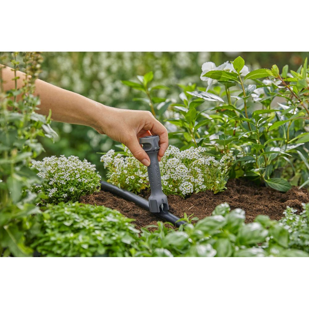 Een hand houdt een grijs gereedschap dat op een zwarte irrigatiebuis in een tuin wordt geplaatst.