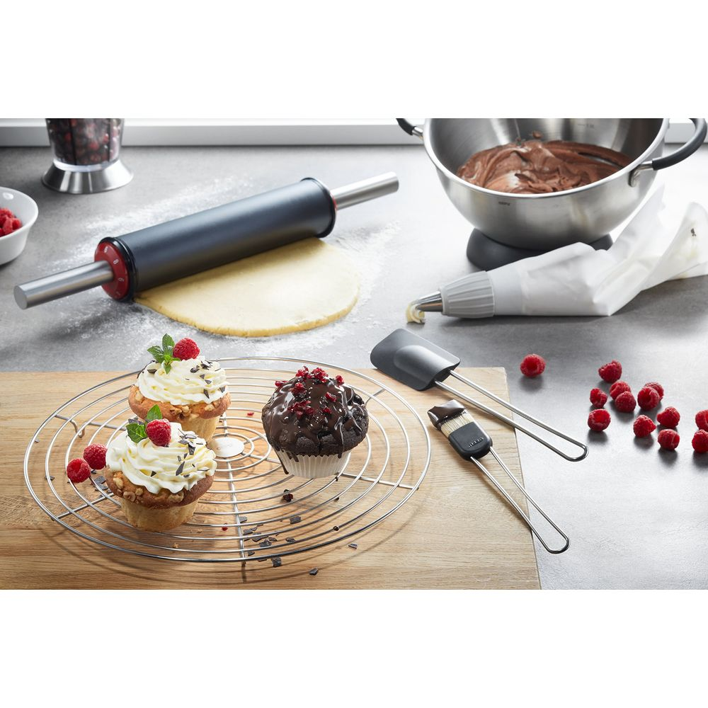 Zwei verzierte Muffins auf einem Rost, ein dritter mit Schokoladenglasur. Backutensilien liegen daneben.