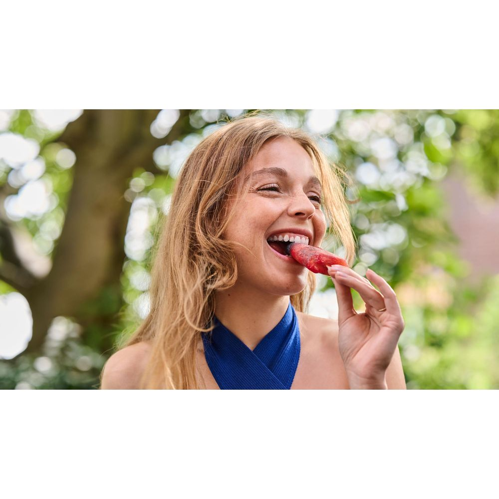 A woman eats a red popsicle outdoors with trees in the background.