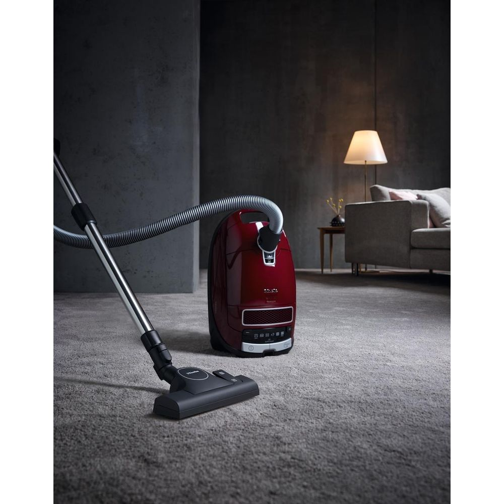 Ein roter Staubsauger steht auf einem grauen Teppich mit einem Schlauch und einer Bodendüse.