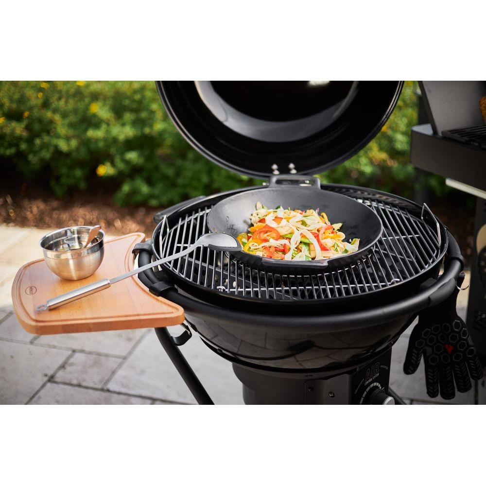Ein Grill mit offenem Deckel, darauf ein Wok mit Gemüse und eine Schöpfkelle.