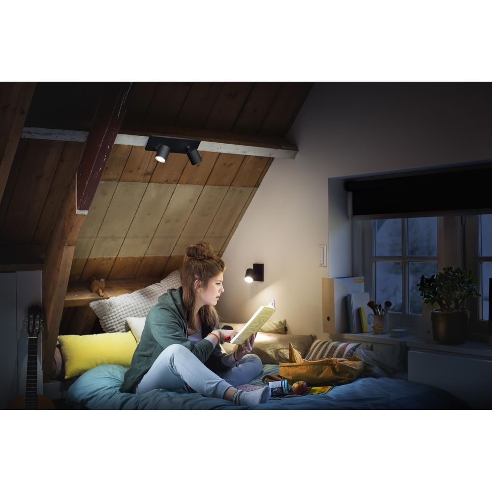 Jonge vrouw leest op bed onder schuine plafonds, verlicht door spots.