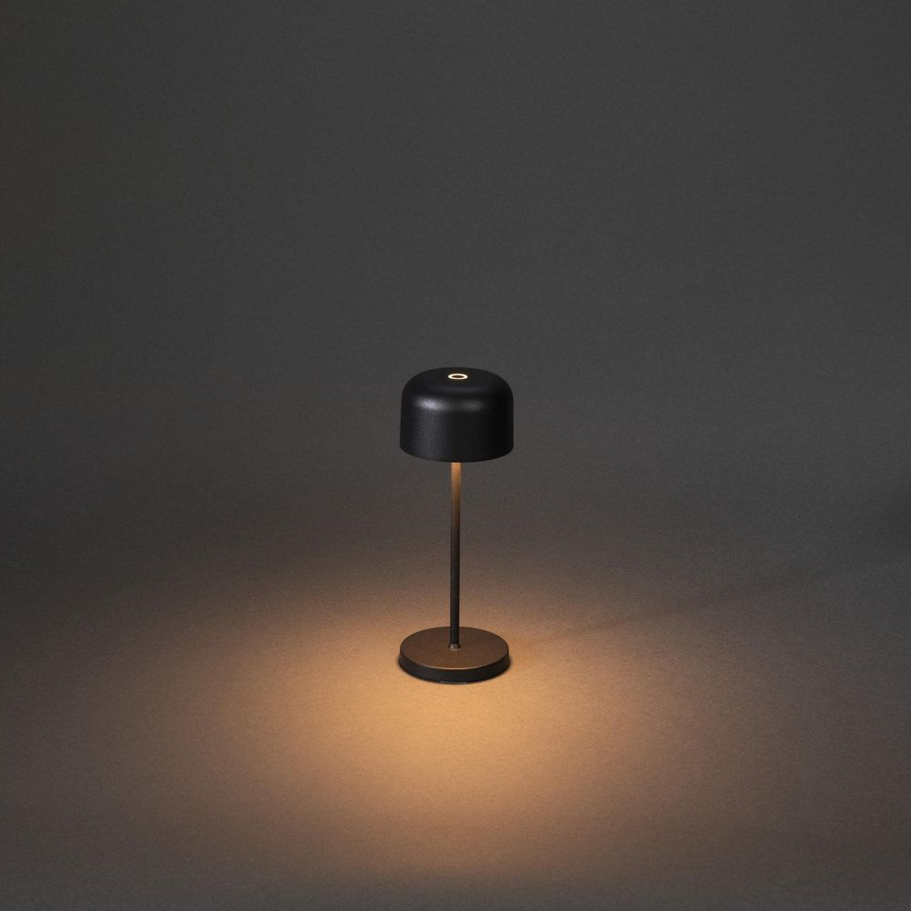 Eine kleine schwarze Tischlampe mit rundem Fuß steht auf einem dunklen Untergrund.