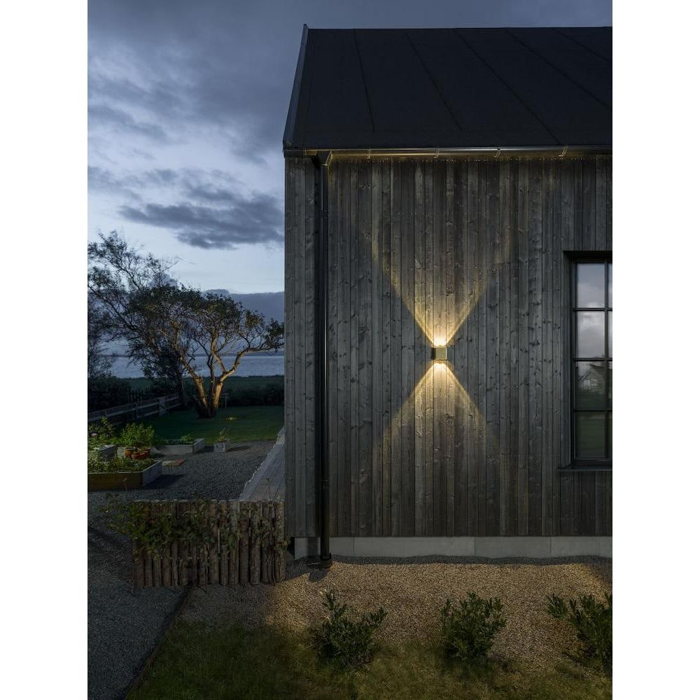 Holzhauswand mit Außenleuchte, die Licht nach oben und unten wirft. Draußen ist es dämmerig.