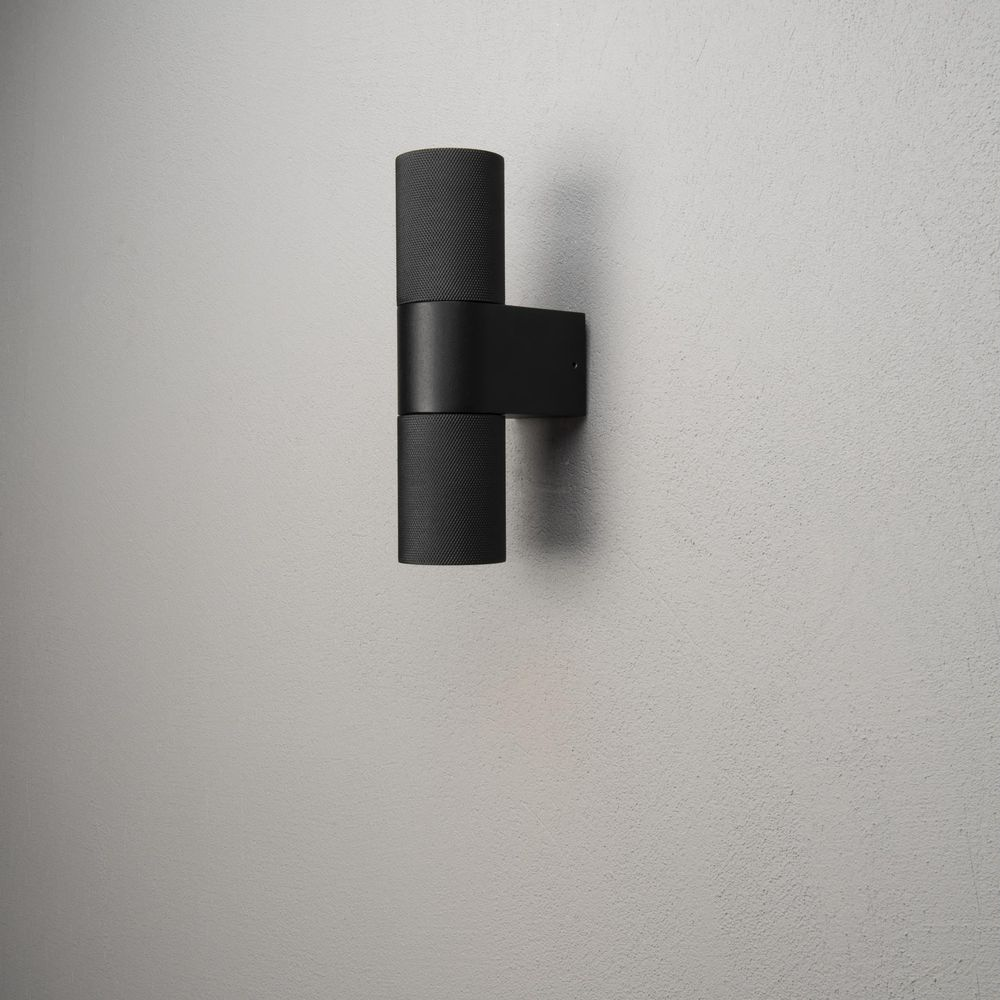 Schwarzer zylindrischer Wandleuchter mit geriffeltem Muster an einer grauen Wand.