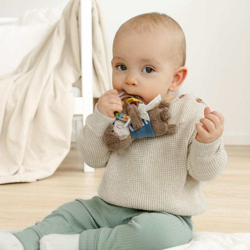 Un bébé est assis par terre jouant avec un anneau de dentition en peluche. Il porte un pull et des leggings.