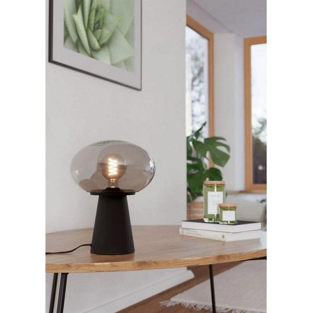 Eine schwarze Lampe mit einem Rauchglas-Schirm steht auf einem Holztisch.