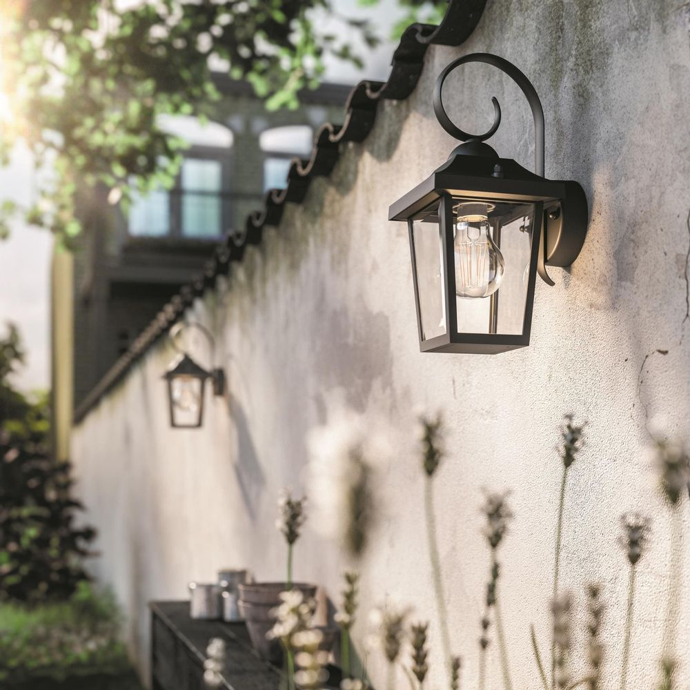Una linterna de pared ilumina un muro blanco con plantas al frente.