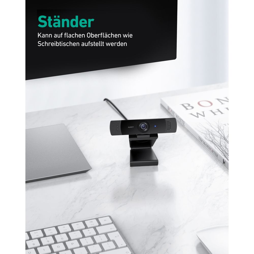 Eine schwarze Webcam mit Ständer steht auf einem Schreibtisch neben einem Laptop und einem Buch.