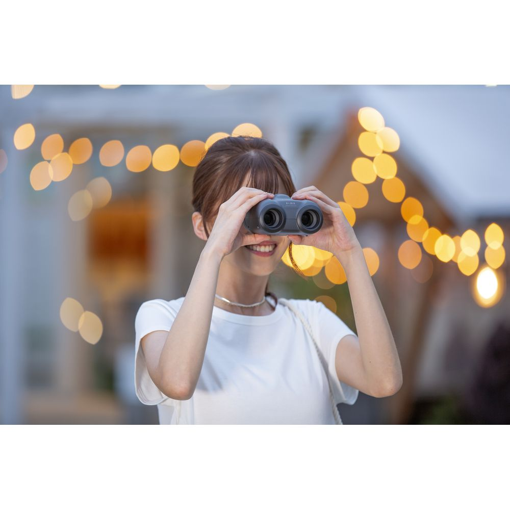 Kobieta w białej koszulce patrzy przez lornetkę, w tle światła.