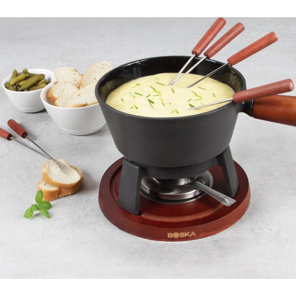 Fondue serowe z chlebem, ogórkami i widelcami do fondue na drewnianym podgrzewaczu.